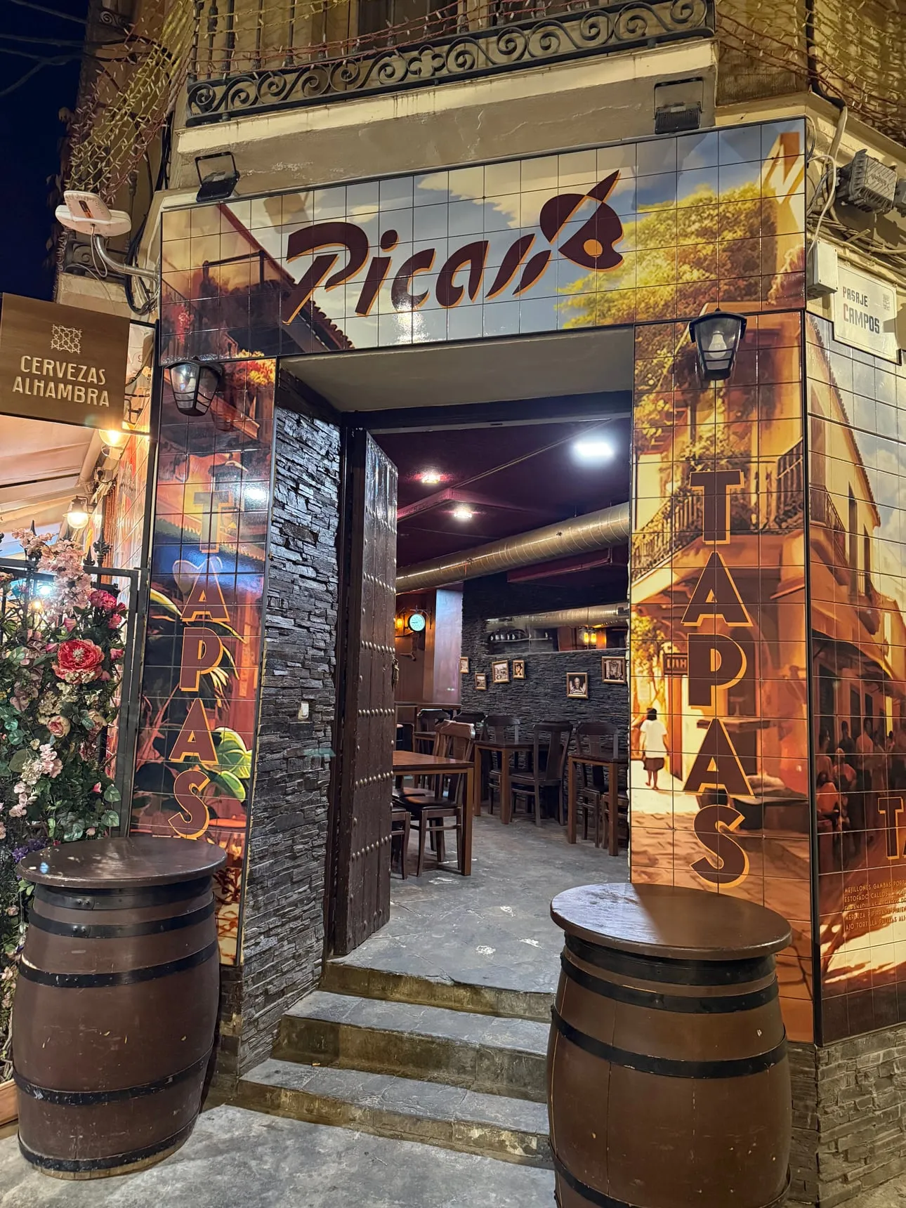 The entrance of Taberna Picas Málaga at night: tile panels with the Picas logo over the door and two barrels on the steps